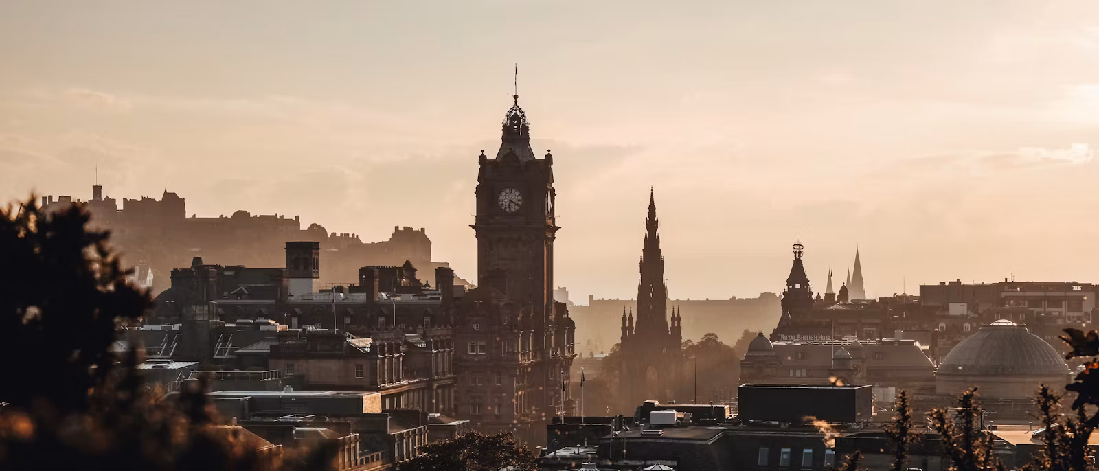The Balmoral Hotel clock tower and the spire of the Scott Monument rising out of hazy golden light over Edinburgh's rooftops, with the castle ridge along the left