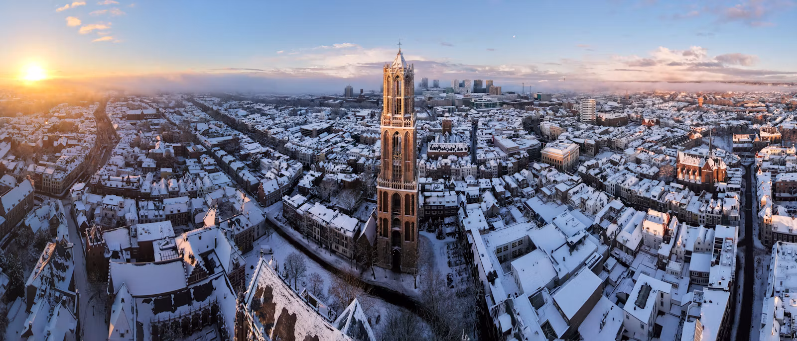 Utrecht from the air at sunrise after snowfall — the Dom tower standing above white roofs in every direction, the sun low on the horizon at the left and the canal a dark line through the old town