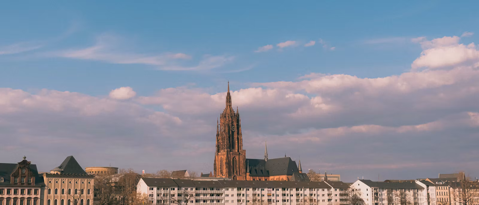 Frankfurt from across the Main — the red sandstone tower of the Kaiserdom rising above the buildings of the north bank under a blue sky with late-afternoon clouds