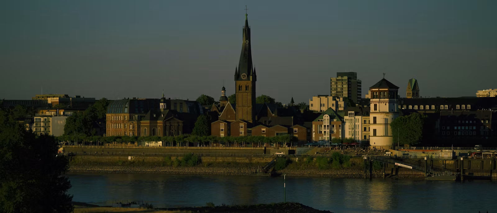 Düsseldorf's Altstadt from the Oberkassel bank of the Rhine in low evening sun — the spire of St. Lambertus and the round Schlossturm above the riverside promenade